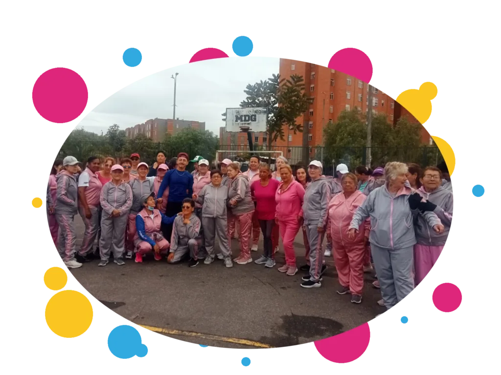 Grupo grande de personas mayores con sudaderas rosadas y grises de la fundación, reunidas junto a la cancha de baloncesto del barrio.