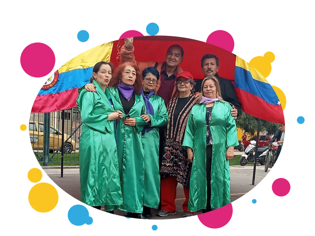Grupo de mujeres mayores con capas verdes y hombres de la comunidad posando frente a banderas de Colombia en una actividad de la fundación.